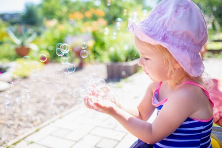 Adorable blonde baby girl 3 year old in a pink hat and blue stripped swimsuit having bath at backyard and playing with bubbles. Summer holidayの写真素材