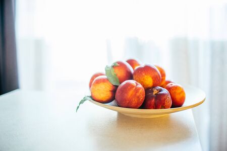 Fresh ripe peaches with leaves on a wooden plate on a kitchen table against the window. Organic food concept and prevention of vitamin deficiency.の写真素材
