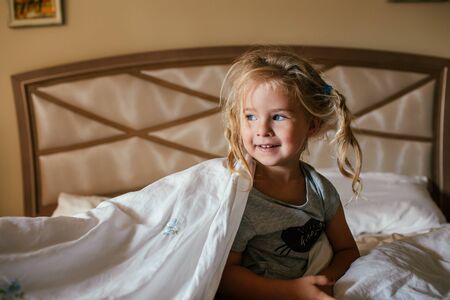 Three years old shaggy white girl sitting on a bed covered with a blanket and smiling.