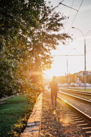 Back view of young sport man running along the railway trucks against the sun in the city. Urban...