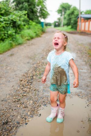 Little blonde girl crying in her dirty clothes after fall into a puddle. Summer, childhood.の写真素材
