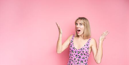 Surprised amazed beautiful blonde woman in pink leopard swimsuit with wide open mouth looking at camera, smiling on a pink backgroundの写真素材
