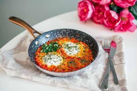Shakshuka with eggs, bell pepper, tomato, and parsley in a cast iron pan on a white table with pink flowers on a background.の写真素材