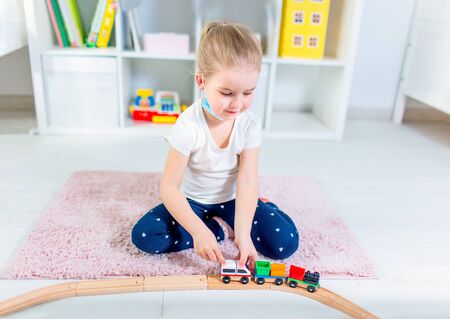 Little blonde girl in a medical mask playing with toy train on a floor in a light room at home during quarantineの写真素材