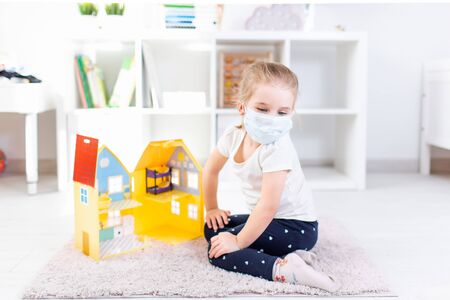 Little blonde girl in a medical mask playing with toy house on a floor in a light room at home during quarantineの写真素材