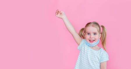 Optimistic, smiling happy little girl taking off the medical mask from face on a pink background with hand up showing end of the pandemia. Copyspaceの写真素材