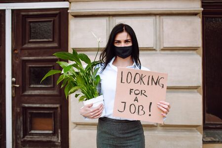 Young business woman holding sign looking for a job and office plant wearing black face mask. Unemployment after corona virusの写真素材