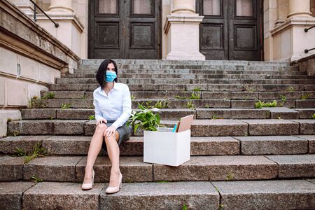 Young business woman in office siut sitting on old stairs with a box of her office supplies and sign looking for a job. Unemployment after corona virusの写真素材