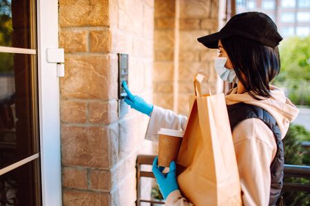 Delivery woman in protective face mask and gloves with a bag of food and coffee ringing with intercom. Health protection, safety and pandemic concept.の写真素材