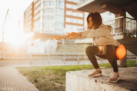 Fit young woman doing squats exercise with fitness gum outdoor in urban environment under the bridge. Slim fit girl stretching outside on a sunsetの写真素材