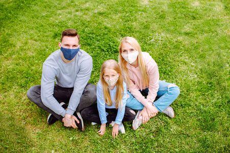 Family members sitting on a grass close each other, smiling in the camera wearing cloth face masks. Father, mother and daughter protect themselves from the virusの写真素材