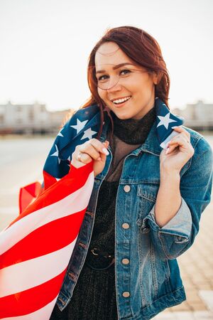Young pretty red haired woman wrapped with national usa flag over sunshine. Soft backlight. Independence day, american dream, freedom conceptの写真素材