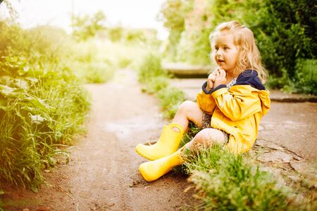 Little blonde girl in yellow raincoat standing and rubber boots sitting on a road after a rain. Summertime, happy childhoodの写真素材