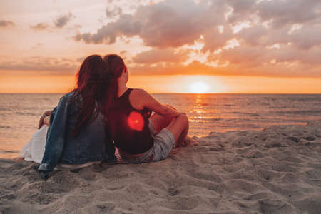 Couple of lesbian girls sitting on the beach and watching and enjoying a beautiful sunset together. Summer time, vacation, LGBTの写真素材