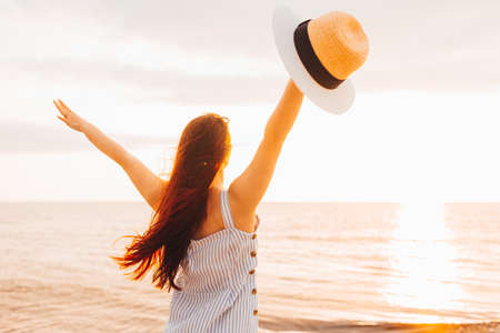 Happy young woman in a dress holding straw hat and and looking at the sunset on empty sand beach with her hands up. Freedoom, vacationの写真素材