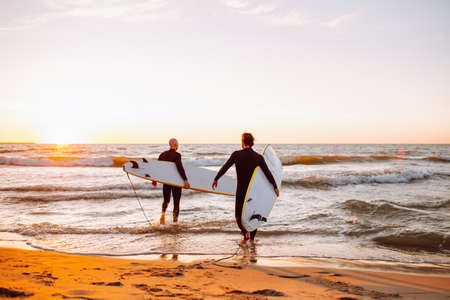 Two young male surfers in black wetsuits with longboards going to water at sunset ocean. Water sport adventure camp and extreme swim on summer vacationの写真素材