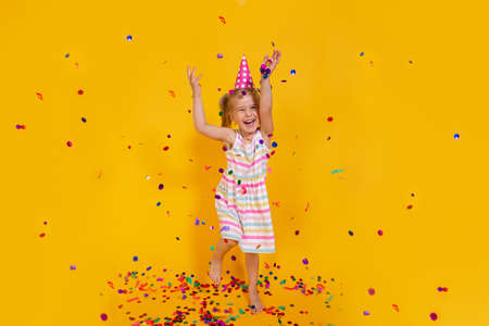 Happy smiling birthday child girl in pink cup surrounded by flying confetti jumping on colored yellow background . Celebration, childhood, emotions.の写真素材