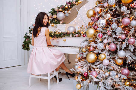 Merry Christmas and New Year. Cheerful womanl in white classic interior playing on a white piano against the background of a decorated Christmas tree.の写真素材