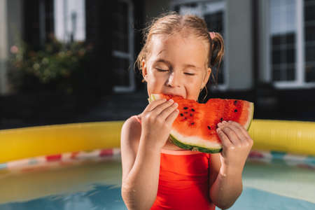 Toddler eating watermelon in pool in the yard with her eyes closed. Kids eat fruit outdoors. 4 years old girl enjoying watermelon.の写真素材