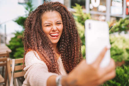Young smiling curly woman takes a selfie with white smartphone sitting at the cafe outside and shows her tongue.の写真素材