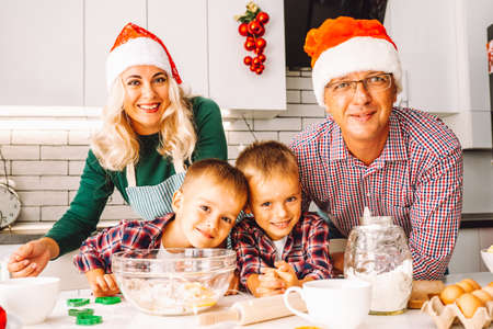Family of two twins boys and age parents preparing cookies for Chtistmas in light kitchen wearing Santa hats and looking at camera.の写真素材