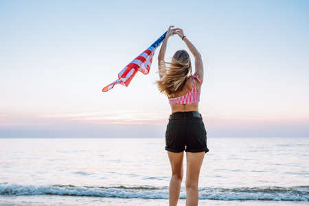 Rear view of unrecognizable young woman holding american waving flag at coastline against of sunsetの写真素材