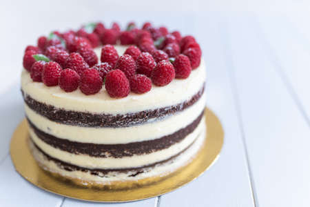 Whole chocolate naked cake with white cheese cream decorated fresh raspberries on a white wooden...