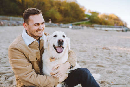 Handsome smiling young man sit on beach and hug his beautiful golden retriever. Trust, care pet conceptの写真素材