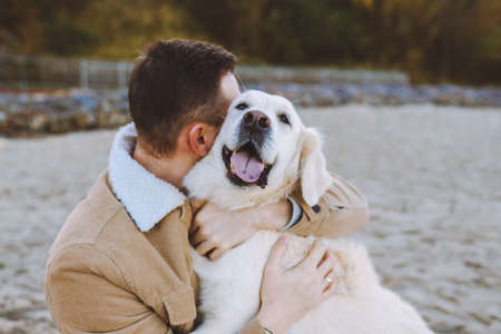 Handsome young man sit on beach and hug and kiss his beautiful golden retriever.の写真素材