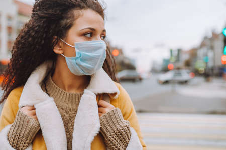 Beautiful afro haired woman wearing protective medical face mask stand on the street of city. Social...