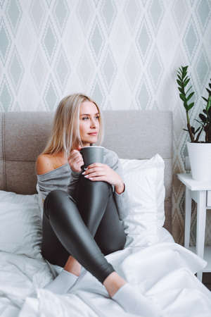 Beautiful blond woman wearing grey home wear sit on bed with cup of coffee at light bedroom and look...