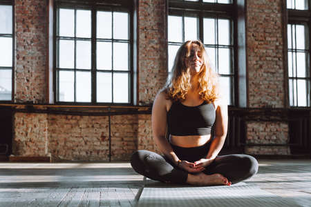 Young curly female sit in lotus pose on floor of loft hall and smile. Positive, meditation, concentrateの写真素材