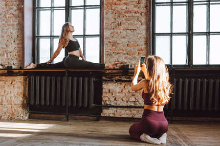 Two female friends take photo after hard workout loft style studio. Woman make picture of her flexible friend making splits on windowsill.の写真素材