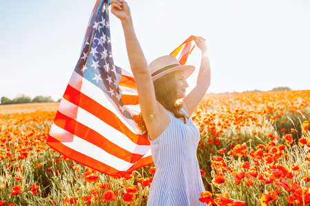 Woman in summer dress and hat with waving flag of United States in poppy field at clear sunny day. Independence Day, National holiday concept.の写真素材