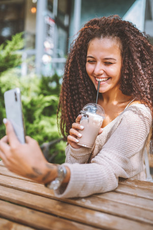 Young smiling afro haired woman take selfie with white smartphone sitting at the cafe outside at sunny summer dayの写真素材