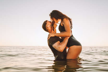 Happy young fit couple embrace each other with love in the sea or ocean at summer sunset. Romantic mood, tenderness, relationship, vacation concept.の写真素材