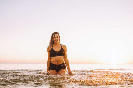 Young sporty woman with beautiful slim body and long dark hair posing in calm sea at sunset in black...