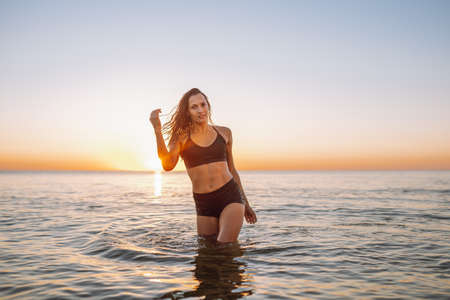 Young sporty woman with beautiful slim body and long dark hair posing in calm sea at sunset in black sportswear. Space for messageの写真素材