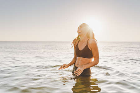 Young sporty woman with beautiful slim body and long dark hair posing in calm sea at sunset in black sportswear. Space for message.の写真素材