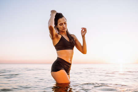 Young sporty woman with beautiful slim body and long dark hair posing in calm sea at sunset in black sportswear. Space for messageの写真素材