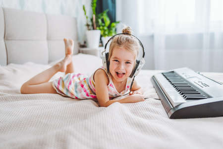 Little blonde toddler girl play classic digital piano at home lying on bed. Home education concept, quarantineの写真素材