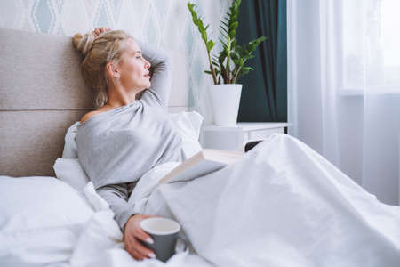 Side view of attractive blonde woman sitting on her bed in light modern bedroom and reading a book with cup of hot beverageの写真素材