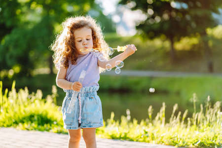 Little curly girl blowing soap bubbles at warm summer day in the parkの写真素材