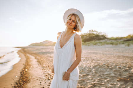 Happy young woman wearing dress and straw hat walk alone along sand beach at sunset along sea shore and smiling.の写真素材