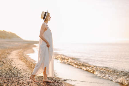 Young slim woman wearing white long dress and hat walk alone at the beach or ocean or sea. Copyspace.の写真素材