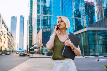 Young business woman standing with laptop among the skyscrapers and speaks on the phone.の写真素材