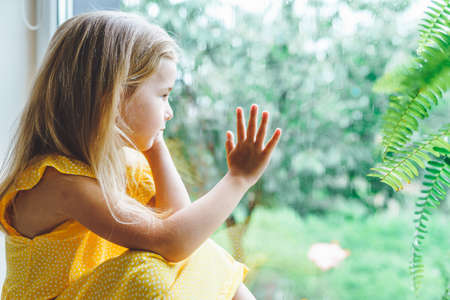 5 years old blonde girl staring out of the window on a wet cold rainy day.