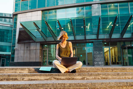 Blonde business woman wearing glasses sit with laptop among the skyscrapers and work.