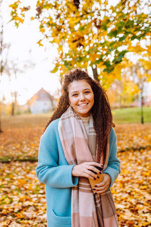 Happy attractive afro-haired woman walk in autumn park at sunny warm day with cup of coffeeの写真素材