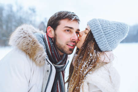 Young happy couple kiss each other and walk in winter park holding hands. Man and woman have fun together during winter holidays.の写真素材